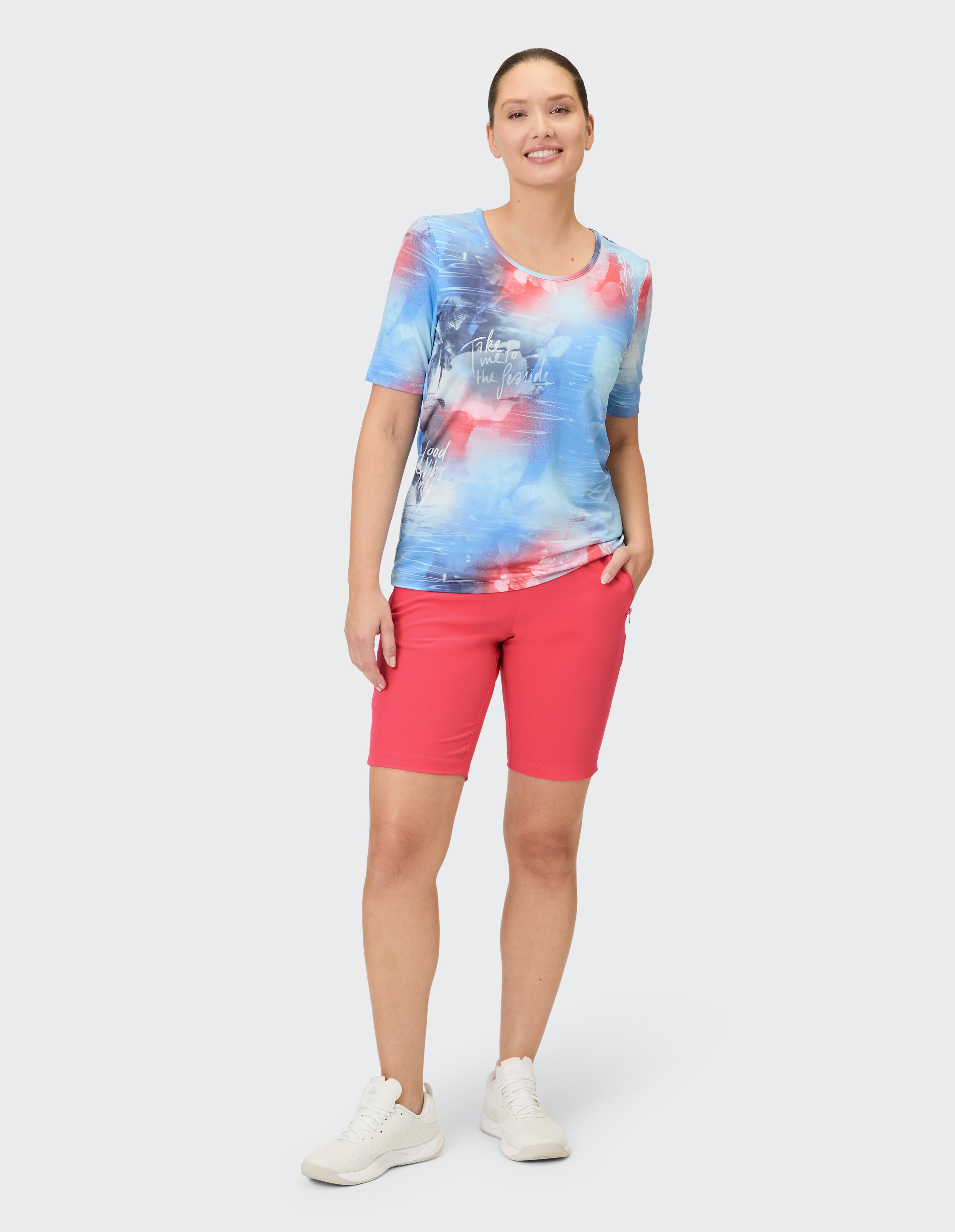 Eine Frau trägt ein kurzärmeliges T-Shirt mit einem blauen, roten und weißen batikartigen Aufdruck und dazu passende rote Bermudashorts.