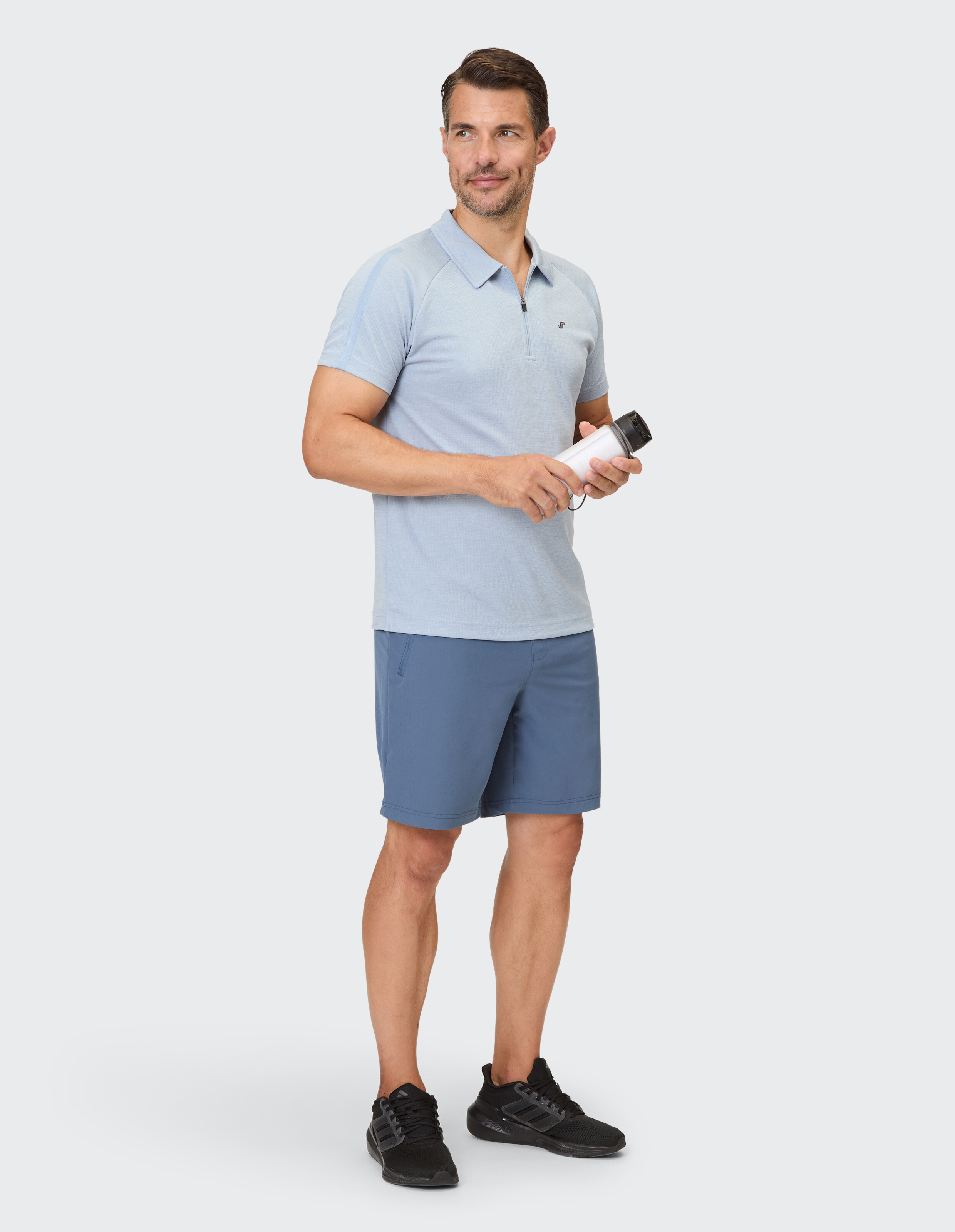 Mann in hellblauem Polo und blauen Shorts, mit einer Wasserflasche in der Hand, lässiger Stil.