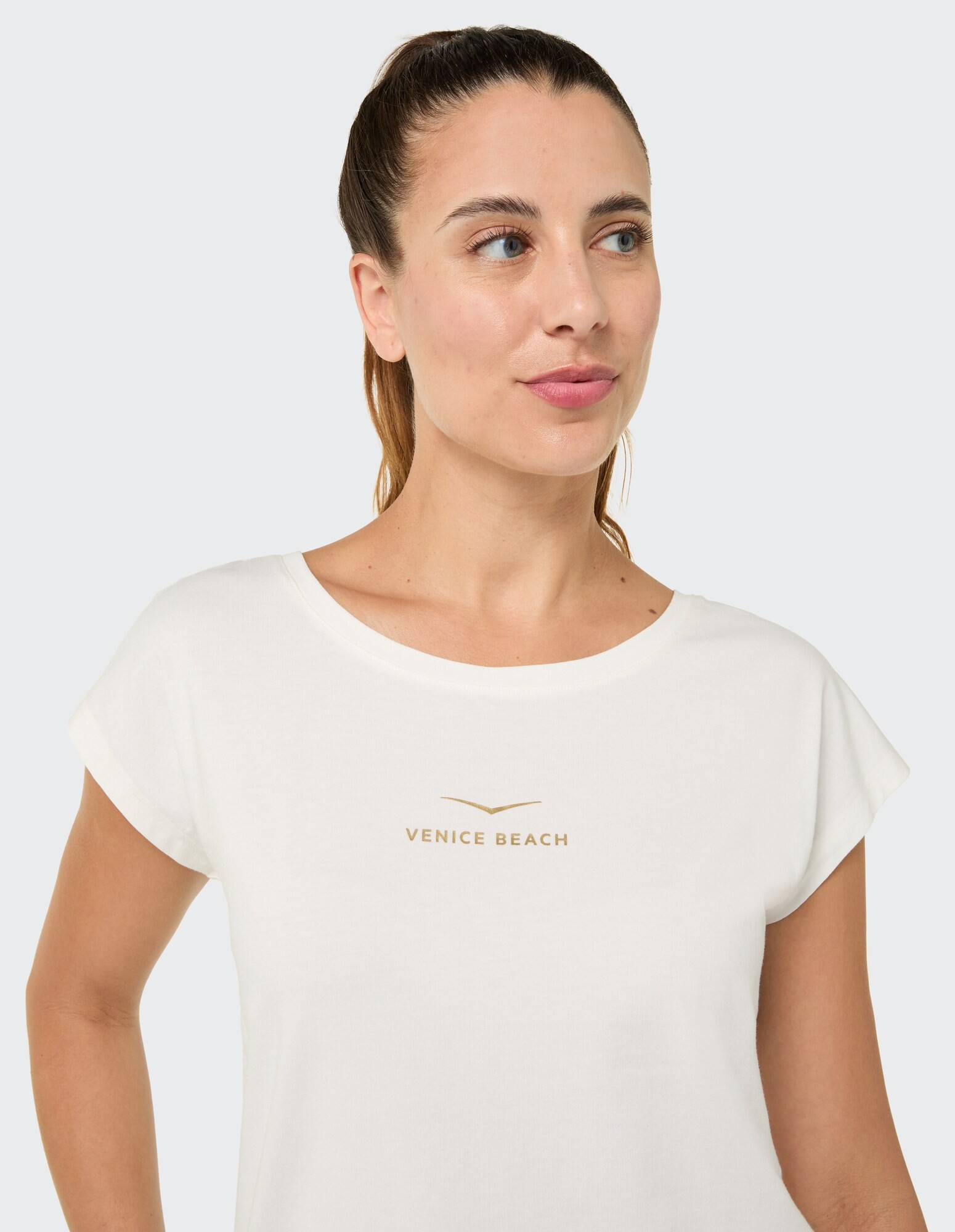 Frau trägt ein weißes Venice Beach-T-Shirt mit lockerer Passform und goldenem Logo.
