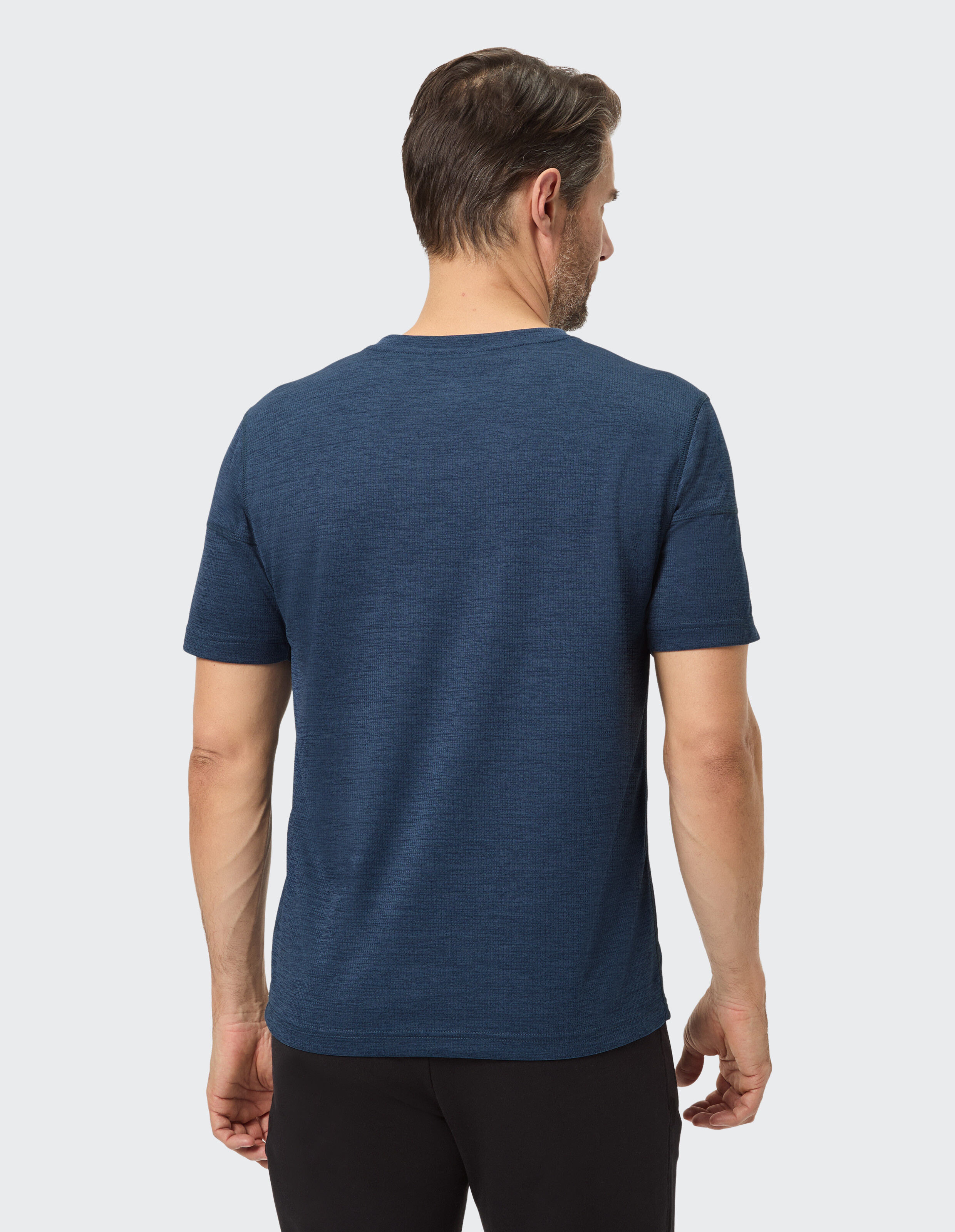 Ein Mann mit braunem Haar trägt ein dunkelblaues T-Shirt und schwarze Hosen, von hinten betrachtet vor einem hellgrauen Hintergrund.