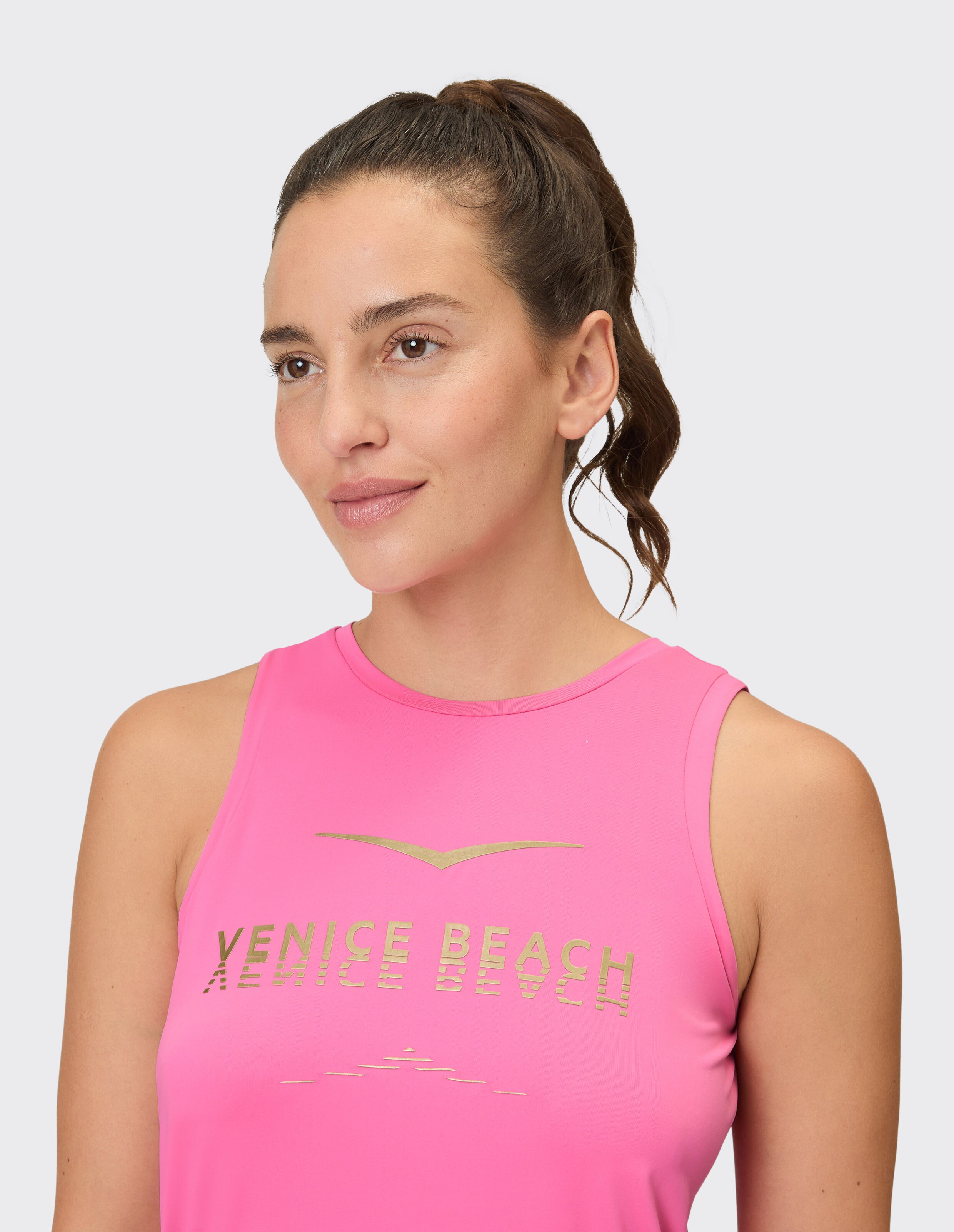 Frau trägt ein rosafarbenes Venice Beach Mila-Tanktop mit goldenem Logo, perfekt für lässige Aktivkleidung.
