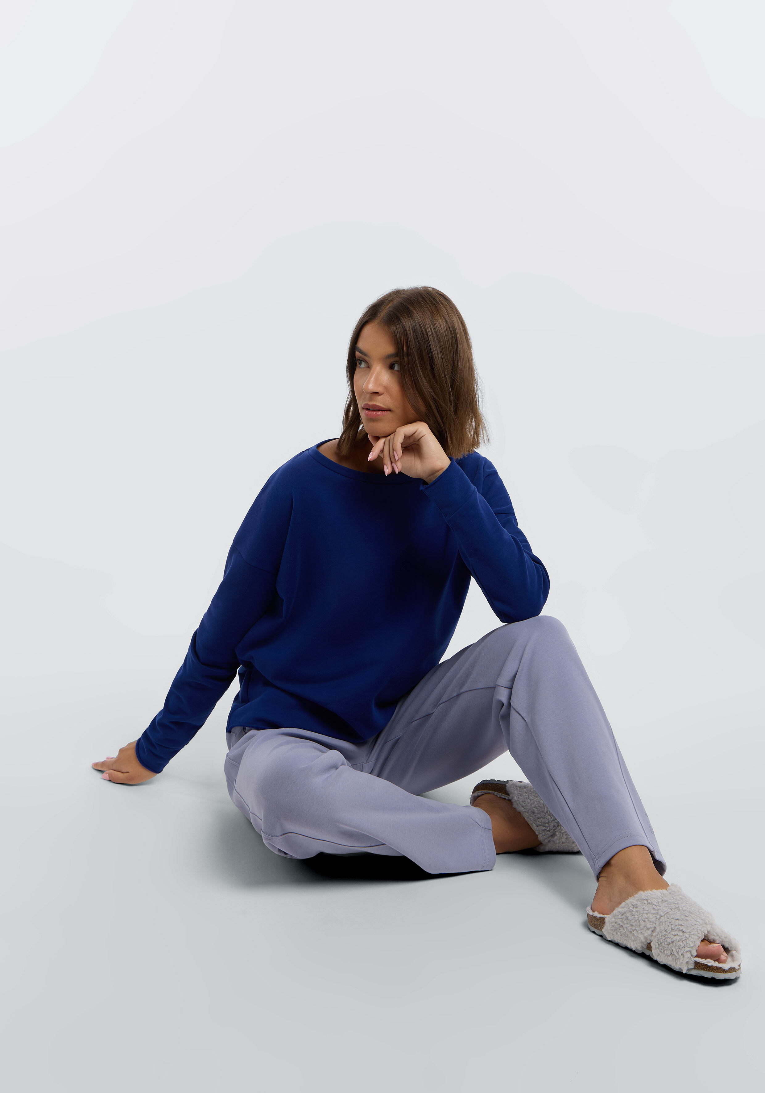 Frau trägt ein blaues Langarmshirt, eine hellviolette Loungehose und flauschige Hausschuhe.