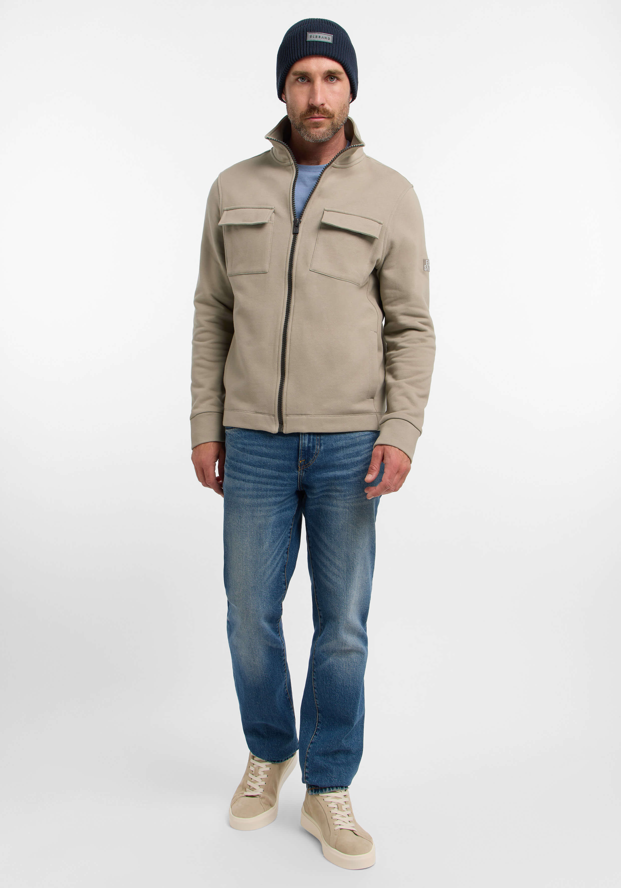 Mann trägt eine beige Jacke mit Reißverschluss, blaue Jeans und eine dunkelblaue Mütze für einen lässigen, gemütlichen Look.