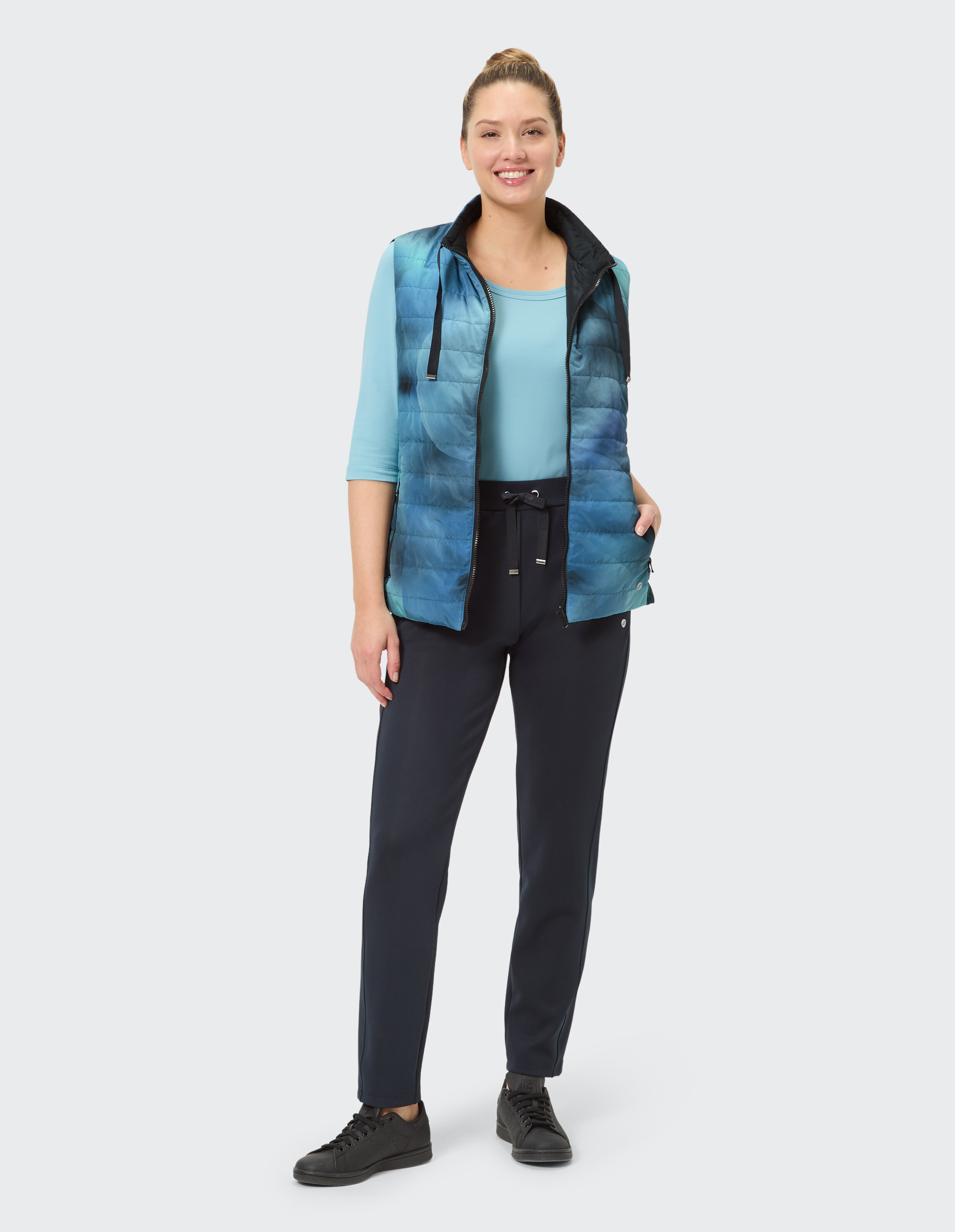 Frau in blauer Weste und hellblauem Langarmshirt, steht lächelnd, trägt dunkle Hose und schwarze Schuhe, neutraler Hintergrund.