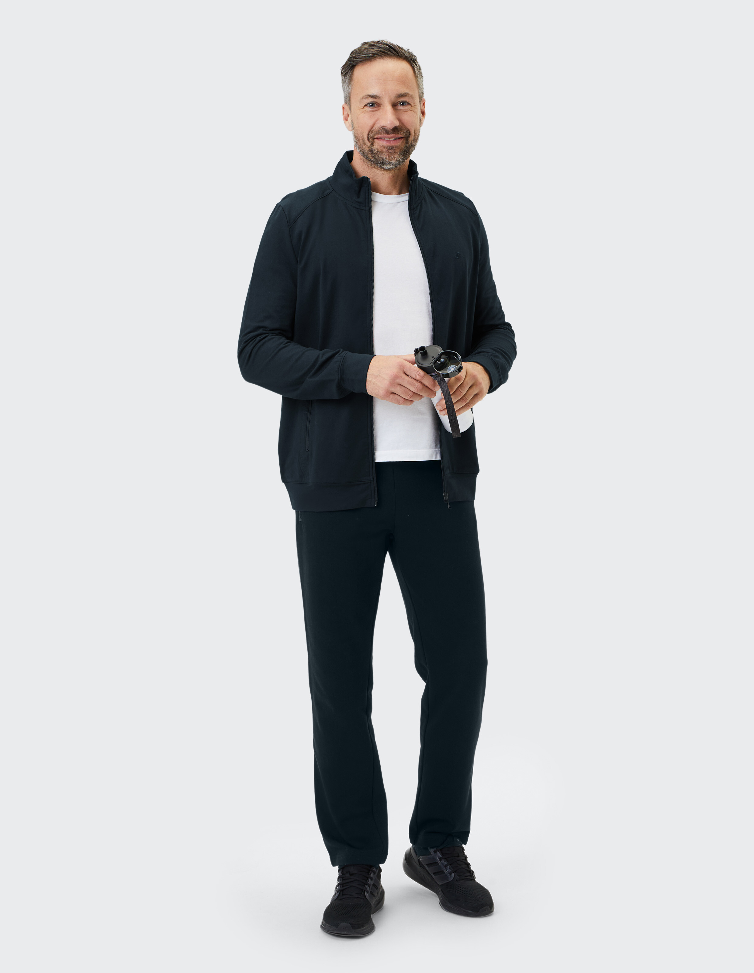 Mann in schwarzer Jacke und Hose mit Reißverschluss, Sonnenbrille in der Hand, lässiger Stil.