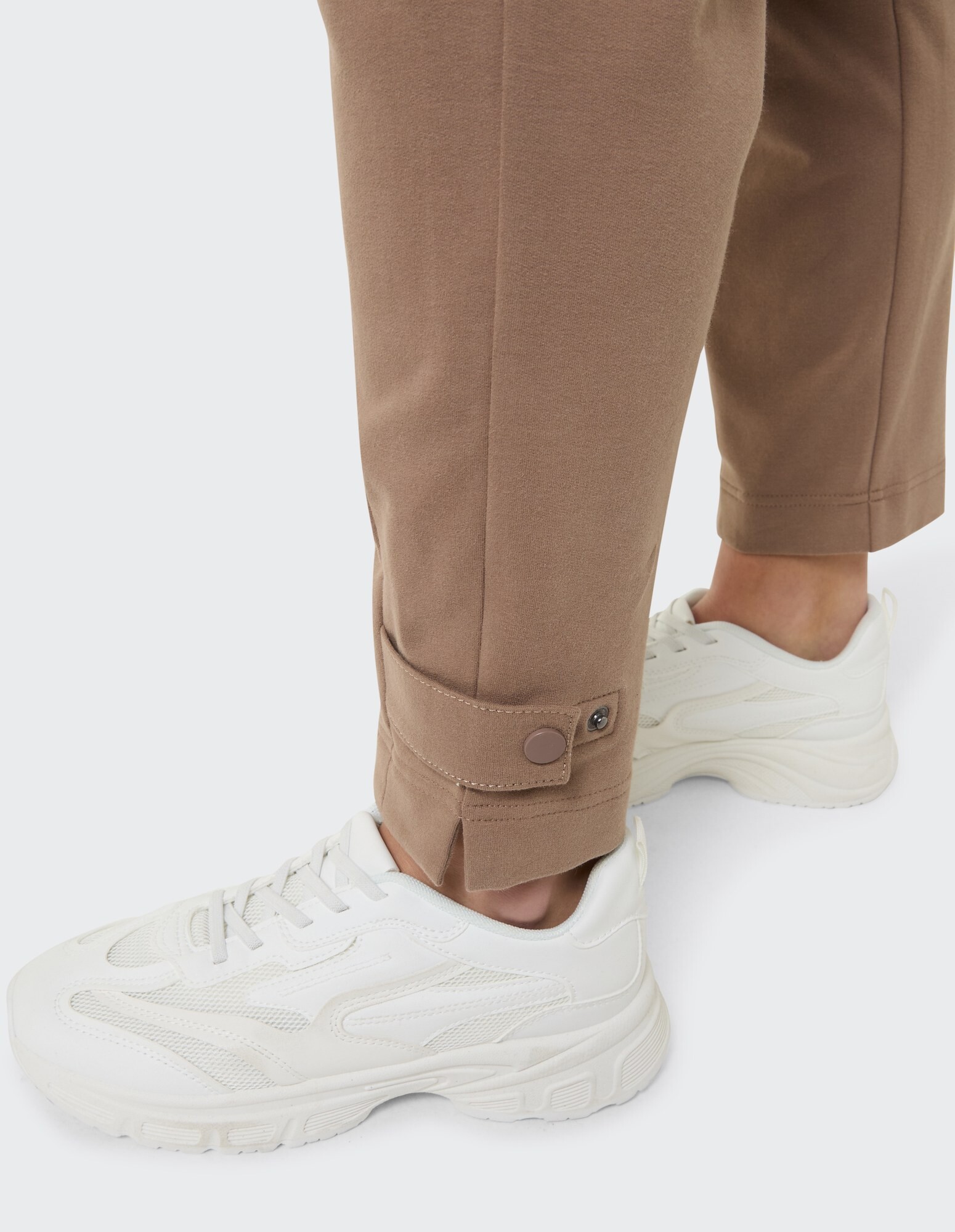Nahaufnahme einer braunen Freizeithose mit Knopfdetail und weißen Turnschuhen.